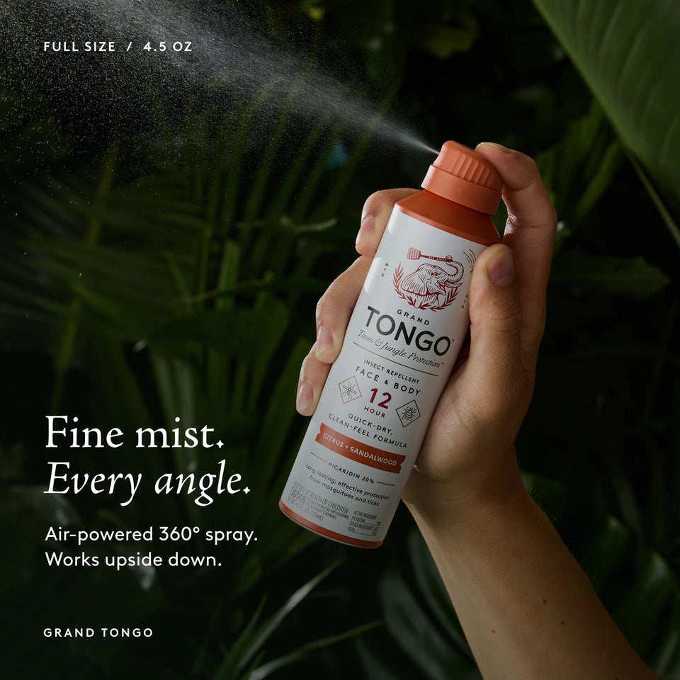 Grand Tongo Citrus + Sandalwood Full Size Spray, 4.5 oz held in a hand, dispensing a visible mist against dark green foliage. Fine mist. Every angle. Air-powered 360° spray. Works upside down.
