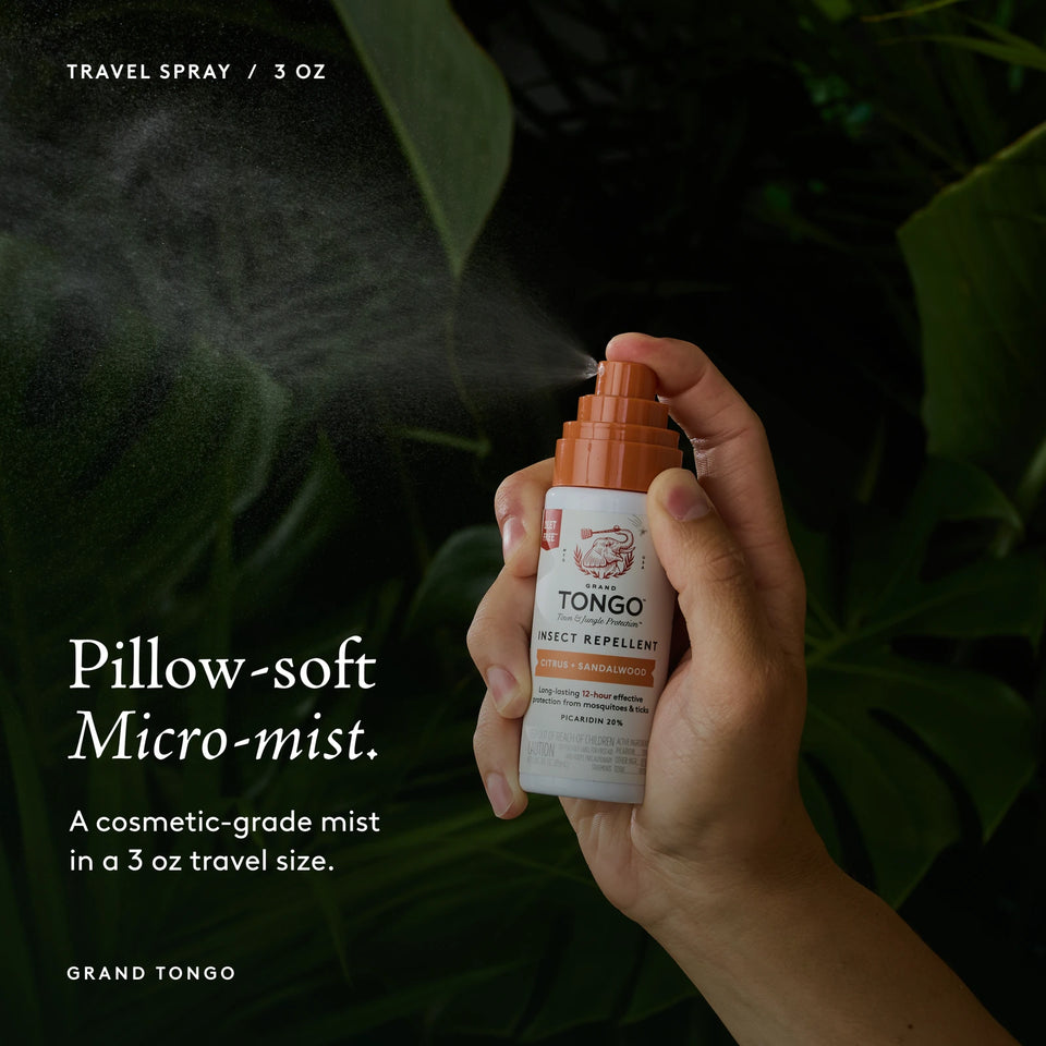 Grand Tongo Citrus + Sandalwood Travel Pump Spray, 3 oz held in a hand, dispensing a visible mist against dark green foliage. Pillow-soft Micro-mist. A cosmetic-grade mist in a 3 oz travel size.