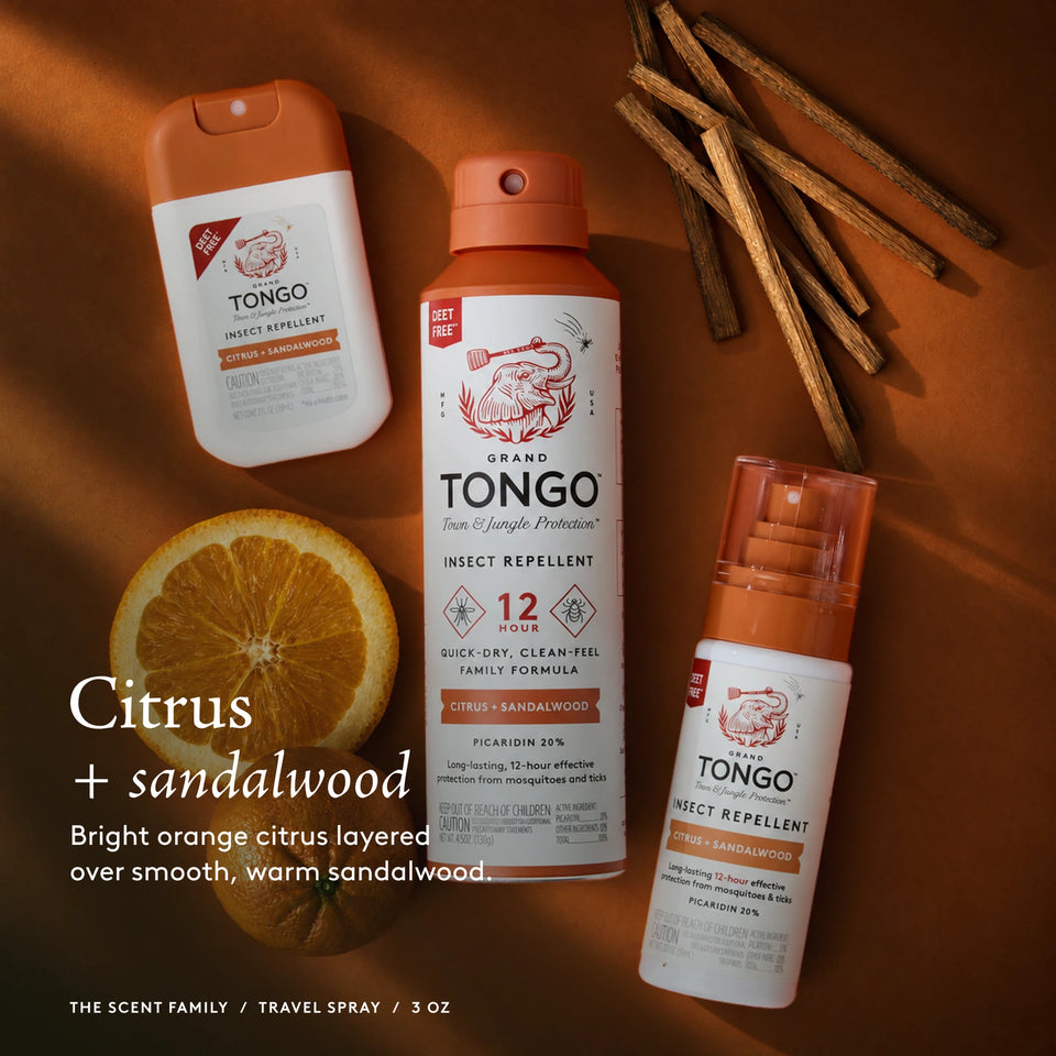 The Grand Tongo Citrus + Sandalwood scent family: full-size, travel, and pocket sprays arranged on orange fabric, citrus, and sandalwood. This is a scent-family image, not the quantity included in the selected product. Citrus + sandalwood. Bright orange citrus layered over smooth, warm sandalwood. The scent family / Travel Pump Spray / 3 oz.